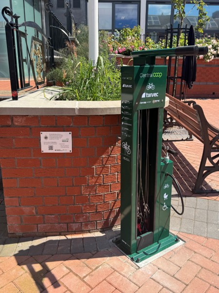 A photo if the bike station in front of the Town Hall, by the railings and a bench with flowers and herbs in the background