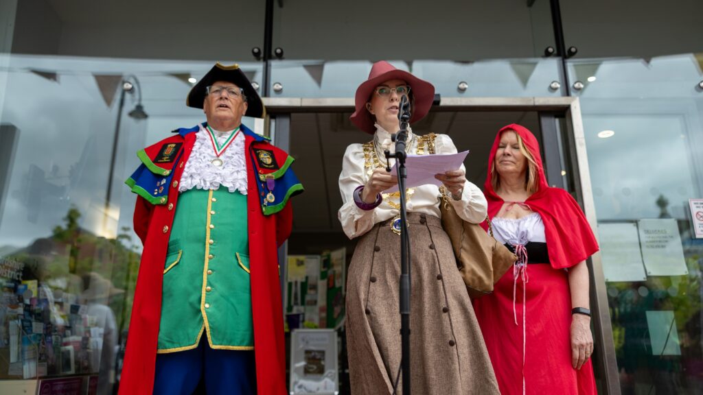 The Mayor, Councillor Charlie Smith, dressed as Miss Marple conducting her speech on the Town Hall steps, the Town Crier is to her right dressed in green, blue and gold and the Festival Organiser, Jackie Neville to her left dressed as Red Riding Hood